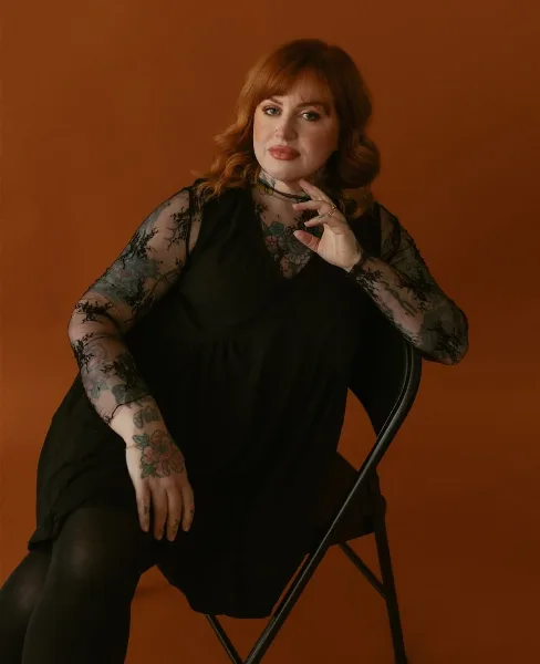 A woman with red hair and tattoos, wearing a black lace top, posing with professional makeup against an orange background.