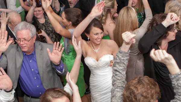 A bride in a white wedding dress dancing on a crowded dance floor surrounded by guests with their hands raised.