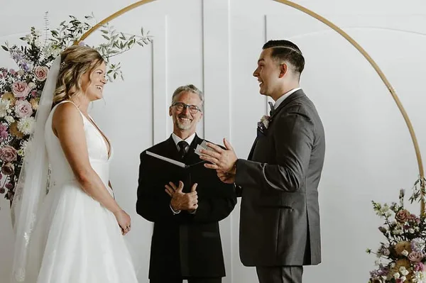 A wedding officiant stands between a bride and groom during a ceremony under a gold arch with floral arrangements.