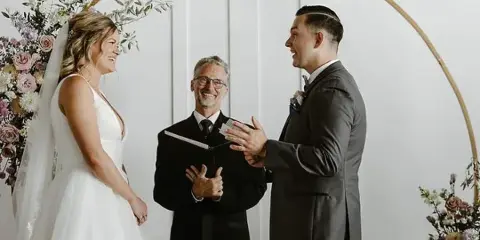 A wedding officiant stands between a bride and groom during a ceremony under a gold arch with floral arrangements.