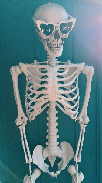 A decorative skeleton wearing heart-shaped sunglasses with wedding initials and a date, set against a teal background.