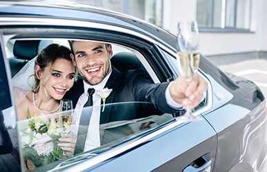 A bride and groom smiling and holding champagne flutes inside a luxury car.