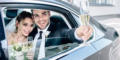 A bride and groom smiling and holding champagne flutes inside a luxury car.