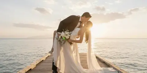 A bride and groom kissing on a wooden pier by the water during a sunset.