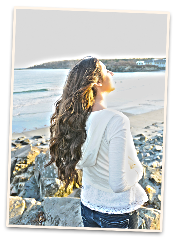 Woman with long styled wavy hair standing on a rocky Maine beach with ocean and shoreline in the background.