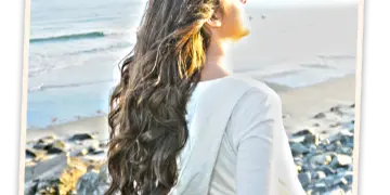 Woman with long styled wavy hair standing on a rocky Maine beach with ocean and shoreline in the background.