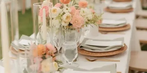 A close-up view of a long wedding reception table featuring a white tablecloth, woven rattan chargers, folded grey napkins, gold cutlery, and a floral centerpiece with pink and white blooms.