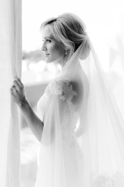 Black and white profile portrait of a bride in a lace dress and veil standing by a window.