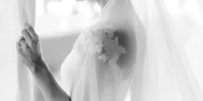 Black and white profile portrait of a bride in a lace dress and veil standing by a window.