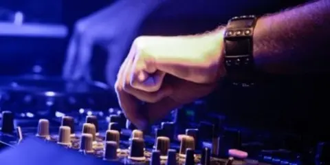 A close-up of a DJ's hands adjusting knobs on a mixing board under blue lighting.