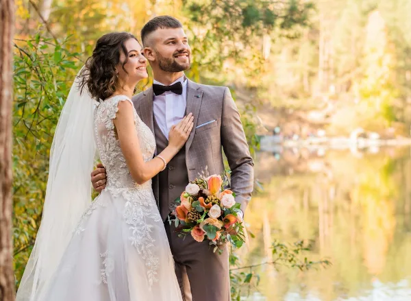 A bride in a white lace gown and a groom in a grey suit standing together outdoors near a body of water.
