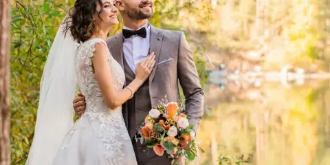 A bride in a white lace gown and a groom in a grey suit standing together outdoors near a body of water.