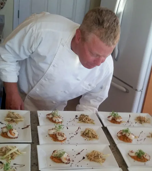 Chef Doug Vaules in a white chef's coat plating a seafood dish with scallops and garnish.
