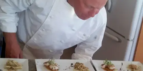 Chef Doug Vaules in a white chef's coat plating a seafood dish with scallops and garnish.