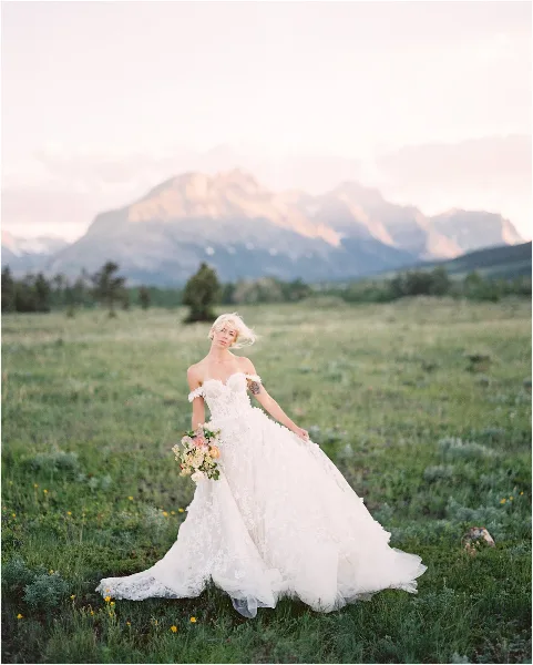 A bride in a white lace wedding dress holding a bouquet, standing in a grassy field with mountains in the background.
