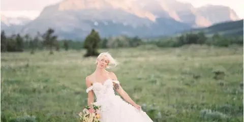 A bride in a white lace wedding dress holding a bouquet, standing in a grassy field with mountains in the background.