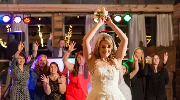 A bride raises her bouquet while dancing with guests at a wedding reception under colorful event lighting.