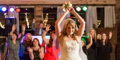A bride raises her bouquet while dancing with guests at a wedding reception under colorful event lighting.