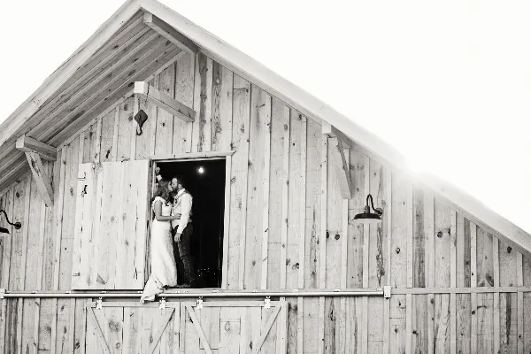 A black and white photograph of a bride and groom standing in the doorway of a rustic wooden barn.