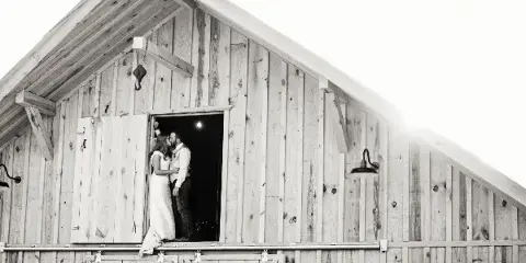 A black and white photograph of a bride and groom standing in the doorway of a rustic wooden barn.