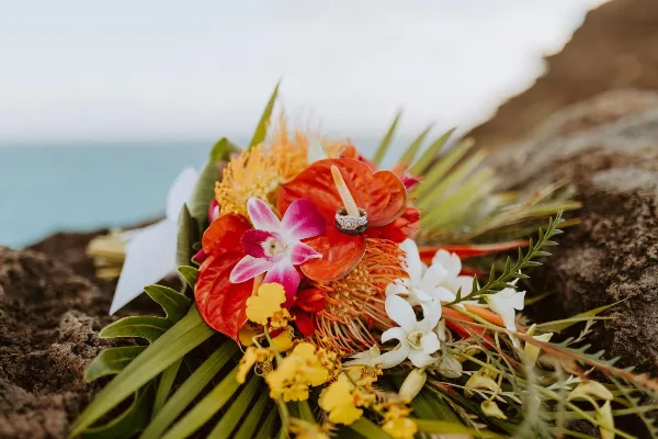 A close-up of a colorful tropical floral arrangement with a wedding ring resting on it, set against a rocky coastal background.