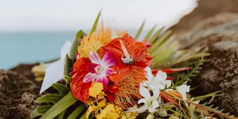 A close-up of a colorful tropical floral arrangement with a wedding ring resting on it, set against a rocky coastal background.