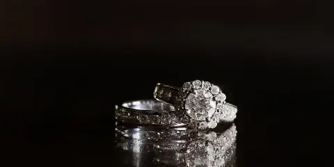 A professional photograph of a diamond engagement ring and a wedding band resting on a dark, reflective surface.