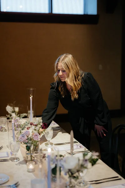 A wedding planner in a black outfit arranging floral centerpieces and table settings at a reception venue.