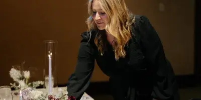 A wedding planner in a black outfit arranging floral centerpieces and table settings at a reception venue.