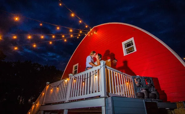 A couple kissing on a white wooden deck in front of a large red barn at night, illuminated by string lights.