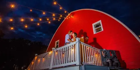 A couple kissing on a white wooden deck in front of a large red barn at night, illuminated by string lights.