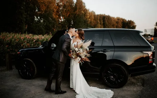 A bride and groom sharing a kiss next to a black luxury SUV.