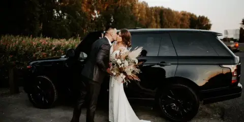 A bride and groom sharing a kiss next to a black luxury SUV.