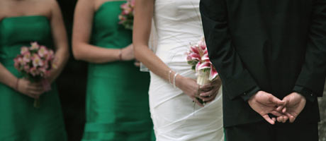 A bride in a white dress and groom in a black suit standing with bridesmaids in green dresses holding bouquets.