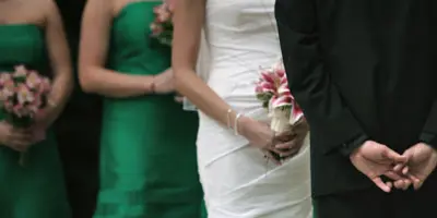 A bride in a white dress and groom in a black suit standing with bridesmaids in green dresses holding bouquets.