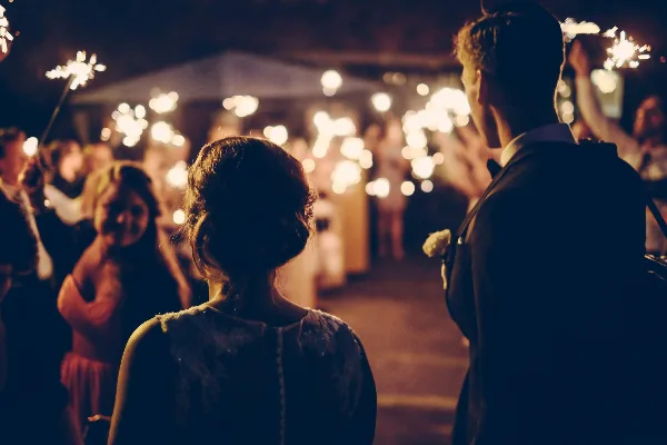A bride and groom at a wedding reception, viewed from behind, with guests holding sparklers in the background.