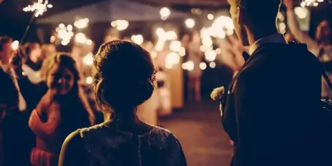 A bride and groom at a wedding reception, viewed from behind, with guests holding sparklers in the background.