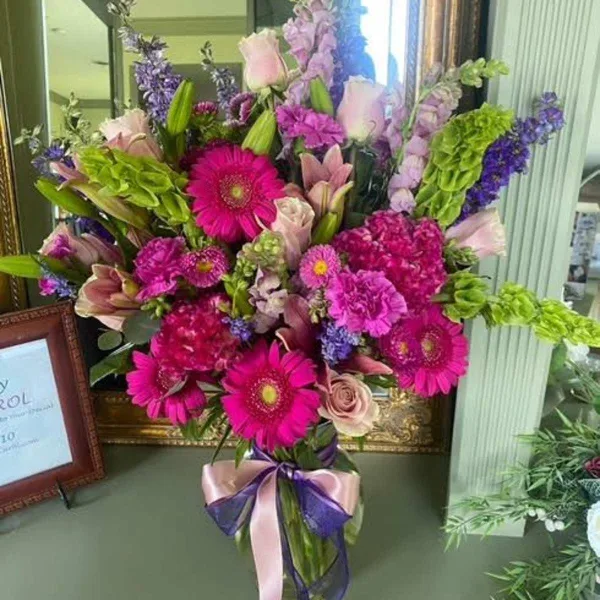 A vibrant floral arrangement featuring pink gerberas, roses, and purple blooms in a vase, displayed in a shop setting.