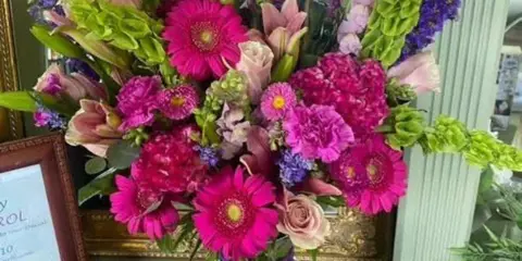 A vibrant floral arrangement featuring pink gerberas, roses, and purple blooms in a vase, displayed in a shop setting.