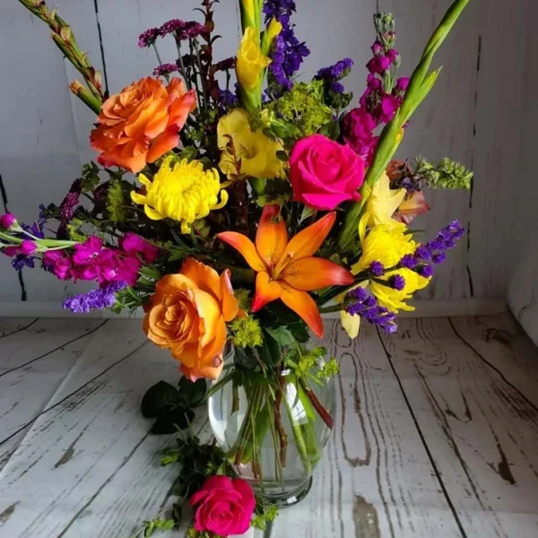 A colorful mixed floral arrangement featuring orange roses, yellow chrysanthemums, and orange lilies in a clear glass vase.