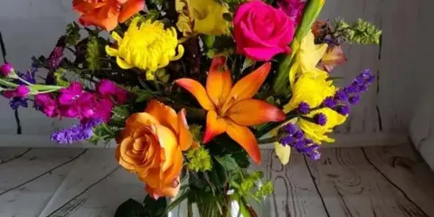 A colorful mixed floral arrangement featuring orange roses, yellow chrysanthemums, and orange lilies in a clear glass vase.