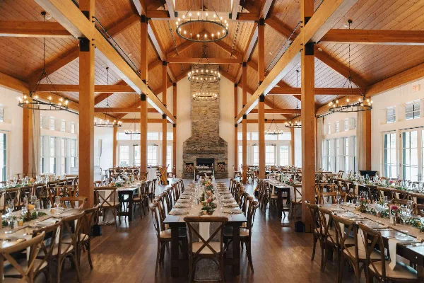 Interior view of a rustic luxury wedding venue featuring exposed wooden beams, a large stone fireplace, and long banquet tables set for a reception.
