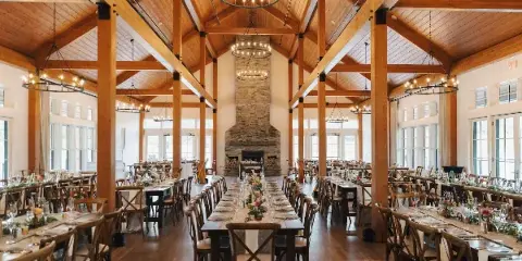 Interior view of a rustic luxury wedding venue featuring exposed wooden beams, a large stone fireplace, and long banquet tables set for a reception.