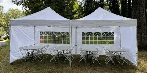 Two white canopy tents set up outdoors on a lawn, furnished with white folding tables and chairs.