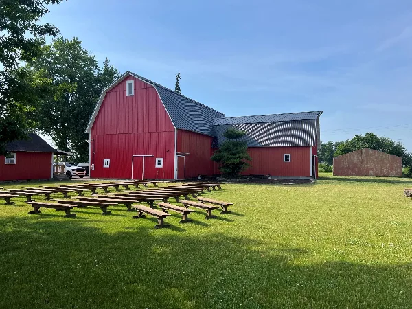 A large red barn with a metal roof and outdoor wooden benches arranged on a green lawn under a clear blue sky.