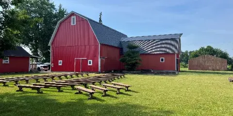 A large red barn with a metal roof and outdoor wooden benches arranged on a green lawn under a clear blue sky.
