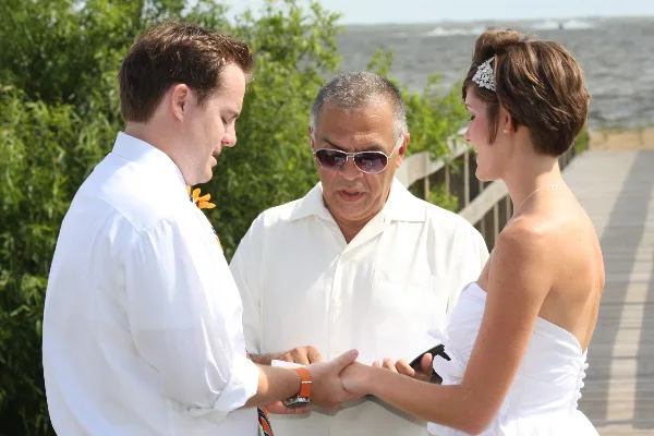 A wedding officiant in a white shirt and sunglasses stands between a bride and groom on a beach, holding their hands during a ceremony.
