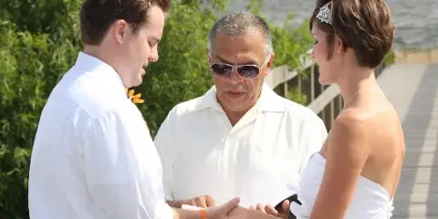 A wedding officiant in a white shirt and sunglasses stands between a bride and groom on a beach, holding their hands during a ceremony.