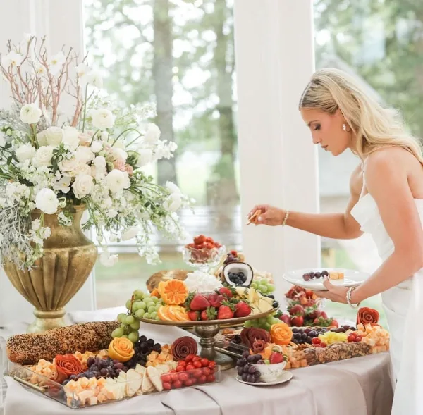 A bride serving herself from an elegant catering table featuring a charcuterie board, fruit platters, and floral arrangements.