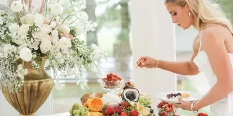 A bride serving herself from an elegant catering table featuring a charcuterie board, fruit platters, and floral arrangements.